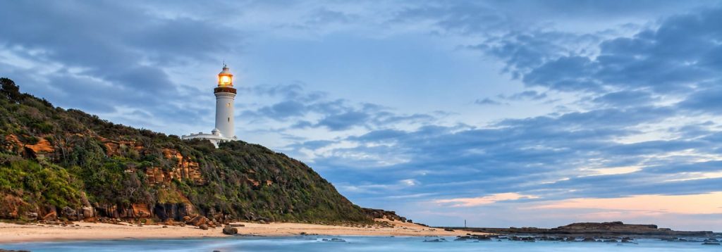 Norah Head Lighthouse glowing at dusk above a rocky shoreline with tide pools